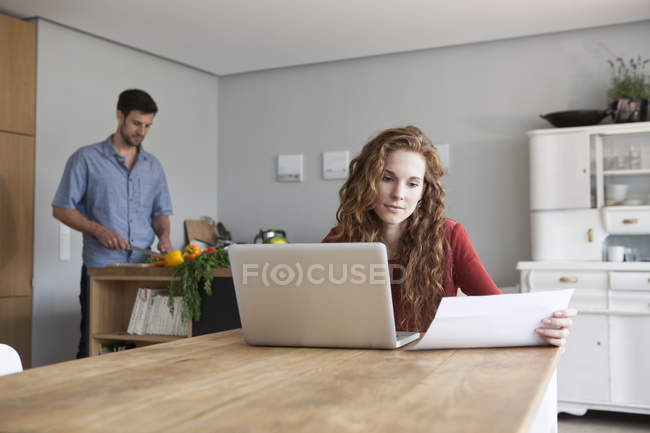Woman at home using laptop with man in background cutting vegetables — Stock Photo