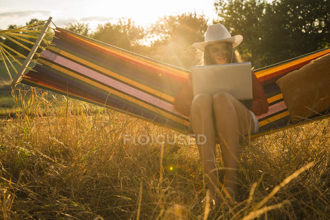 Donna che indossa il cappello seduta su un'amaca usando il computer portatile — Foto stock