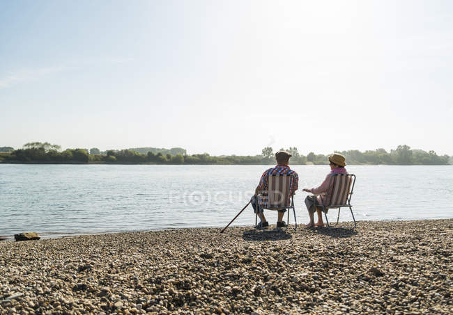 Vista posteriore della coppia anziana seduta fianco a fianco su sedie pieghevoli lungo il fiume — Foto stock