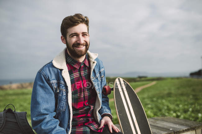 Lächelnder junger Hipster-Mann sitzt mit Longboard auf Bank — Stockfoto