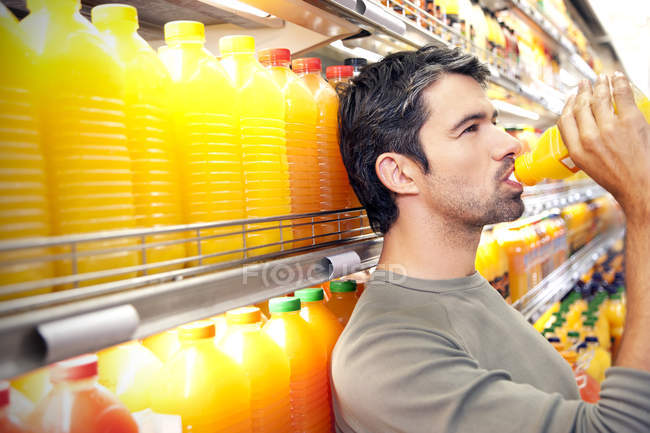 Man drinking a bottle of orange juice  in front of fridge in a supermarket — Stock Photo