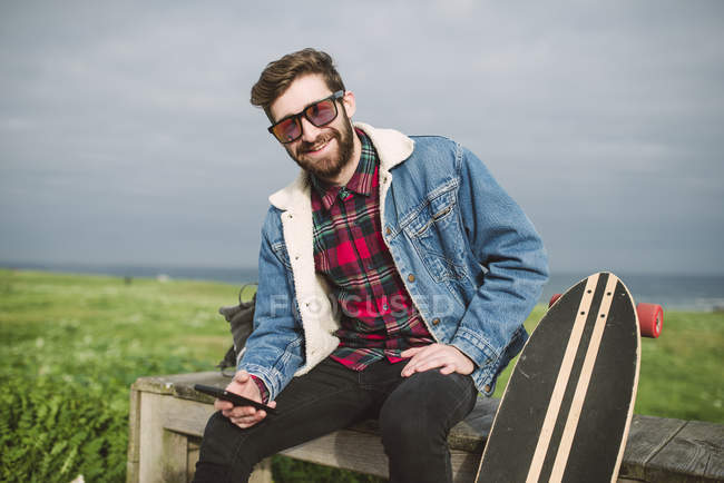 Joven hipster hombre sentado en el parque con longboard y la celebración de teléfono inteligente — Stock Photo