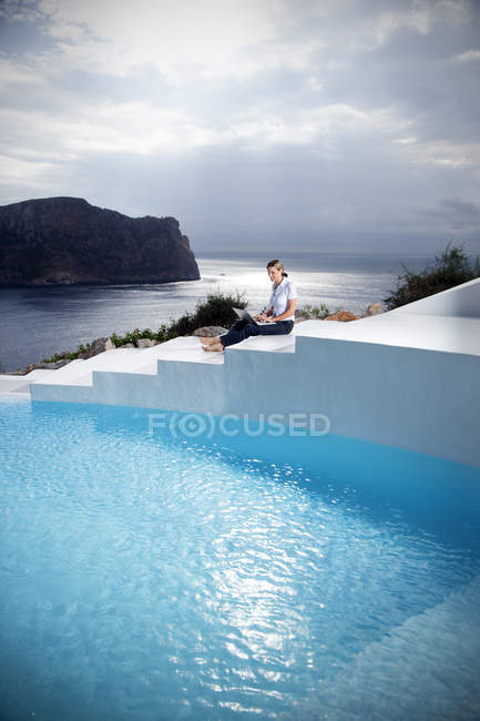 Espanha, Maiorca, mulher com laptop sentado em escadas, além de uma piscina com o mar no fundo — Fotografia de Stock