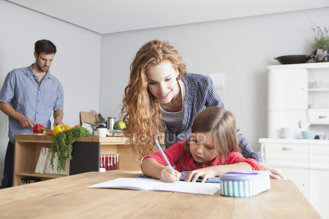 Little girl doing homework at the kitchen table with her parents in the background — Stock Photo