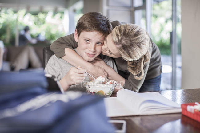 Mother kissing son eating fruit salad and doing homework — Stock Photo