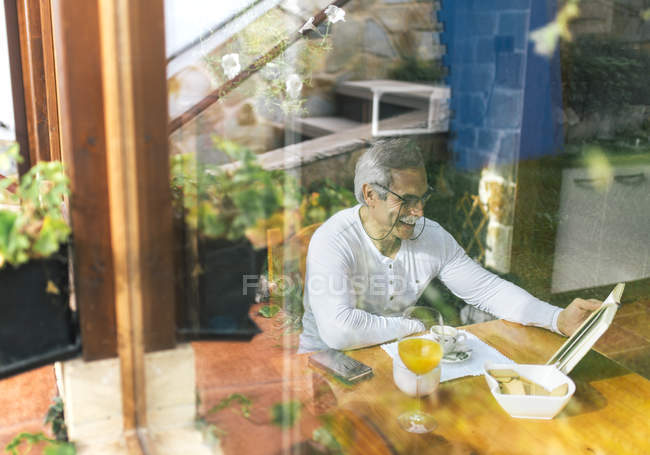 Man behind windowpane sitting at breakfast table reading a book — Stock Photo
