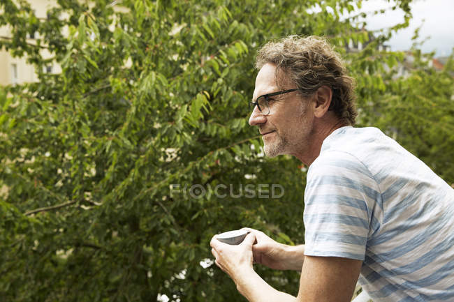 Ältere Mann Seitenansicht auf dem Balkon mit Papier Kaffeetasse, grünes Laub auf dem Hintergrund — Stockfoto