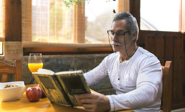 Homme assis à la table du petit déjeuner lisant un livre — Photo de stock
