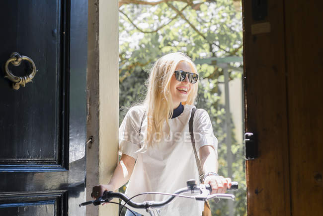 Portrait of laughing blond woman with bicycle standing at opened entrance door — Stock Photo