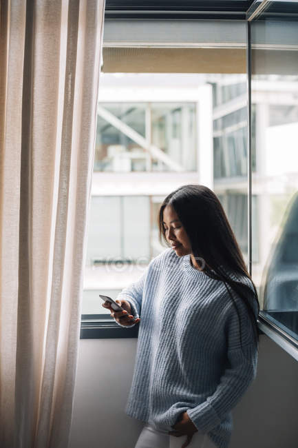 Young woman with cup of coffee standing in front of open window looking at cell phone — Stock Photo