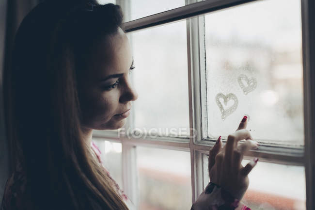 Young woman drawing hearts on fogged-up windowpane — Stock Photo