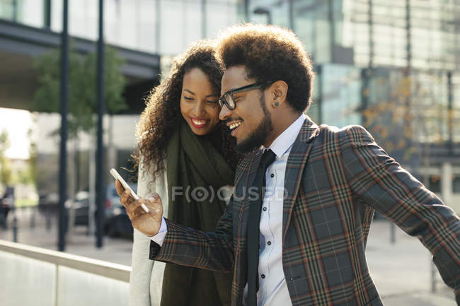 Zwei lächelnde junge Geschäftsleute teilen ihr Smartphone vor einem Bürogebäude — Stockfoto