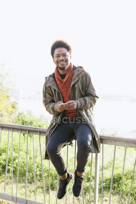 Портрет улыбающегося молодого человека со смартфоном, сидящего на перилах — стоковое фото
