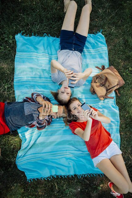 Three teenage friends with smartphones lying on blanket — Stock Photo