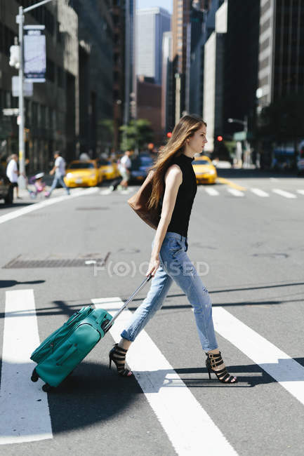 USA, New York City, young woman with rolling suitcase crossing a street — Stock Photo