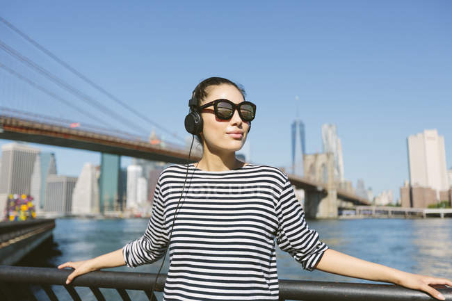 USA, New York City, portrait de jeune femme avec écouteurs et lunettes de soleil devant skyline — Photo de stock