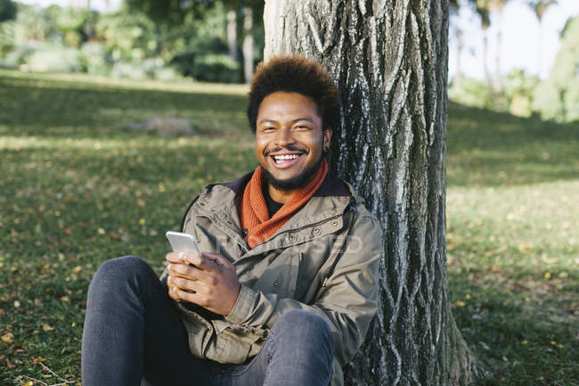 Portrait de jeune homme souriant avec smartphone appuyé contre le tronc d'arbre — Photo de stock
