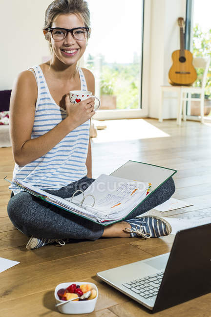 Smiling young woman sitting on wooden floor with file folder and laptop — Stock Photo