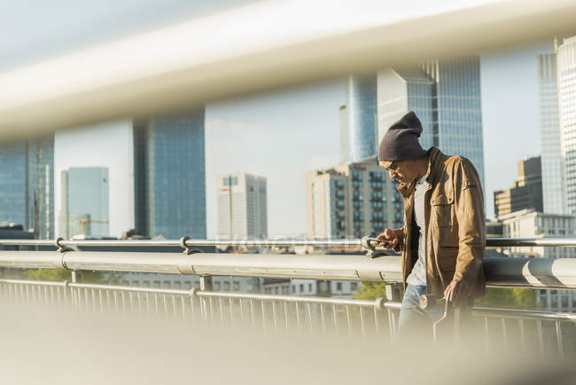 Germany, Frankfurt, man on bridge with skateboard looking on smartphone — Stock Photo