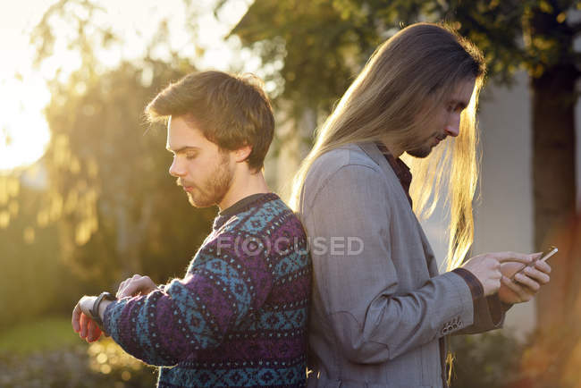 Dos hipsters espalda con espalda con smartwatch y smartphone - foto de stock