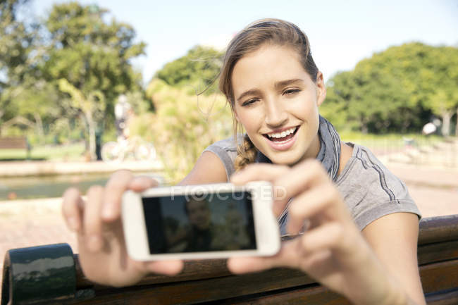 Feliz joven en el banco del parque tomando una selfie - foto de stock