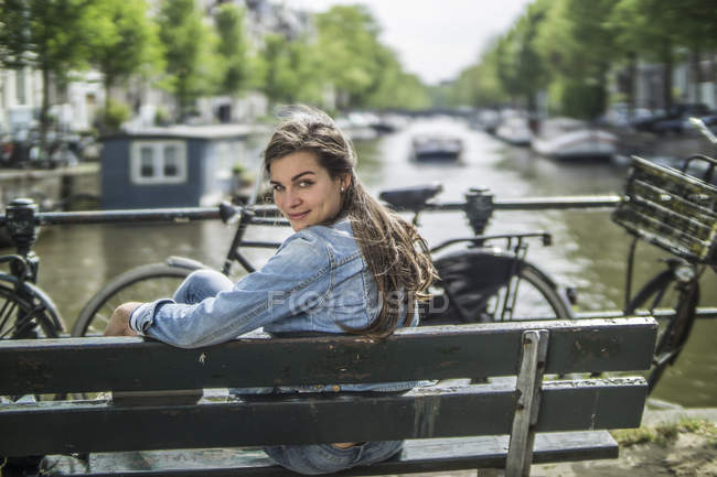 Pays-Bas, Amsterdam, femme souriante assise sur un banc devant le canal de la ville — Photo de stock