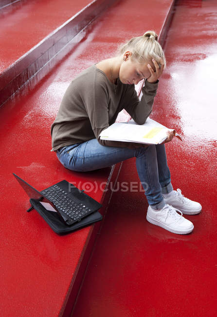 Adolescente femenina con folleto y lectura portátil en escaleras rojas - foto de stock
