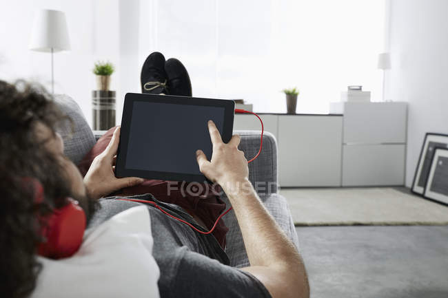 Young man lying on couch at home listening music with headphones and digital tablet — Stock Photo