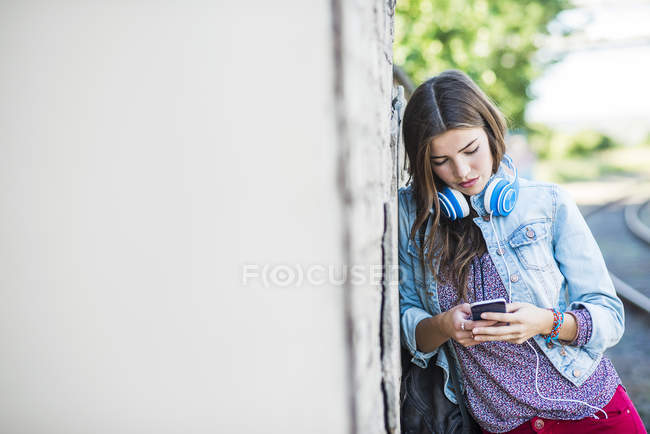 Bruna giovane donna con cuffie e telefono cellulare appoggiato al muro — Foto stock