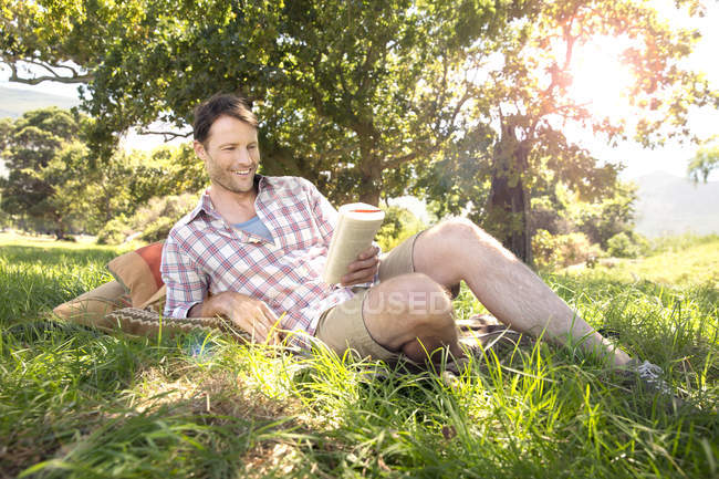 Uomo sorridente sdraiato sul prato lettura libro — Foto stock