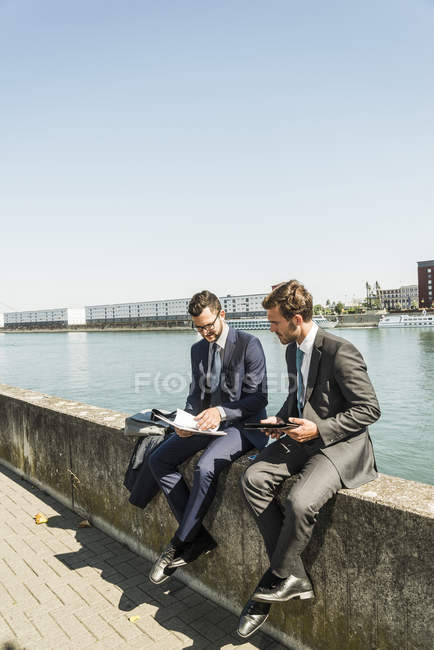Dos jóvenes empresarios sentados en la pared junto al río, trabajando - foto de stock