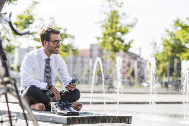 Joven hombre de negocios sentado con las piernas cruzadas en el banco, utilizando el teléfono inteligente - foto de stock