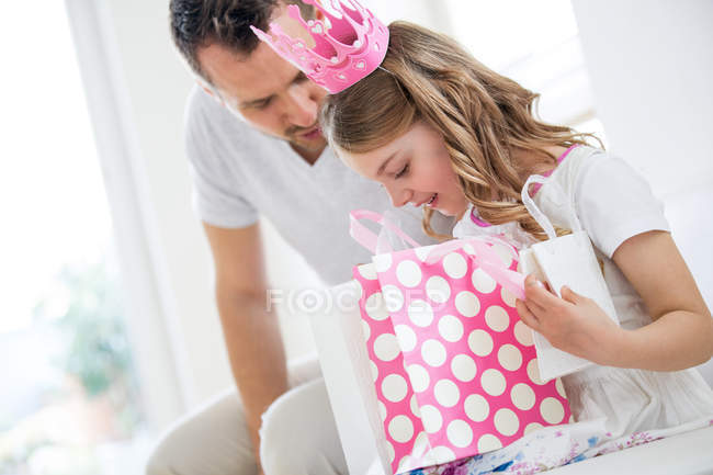 Девушка в розовой короне с подарочными пакетами, отец смотрит дома — стоковое фото