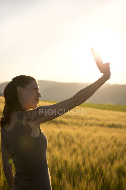 Femme debout devant un champ au lever du soleil prenant un selfie avec son smartphone — Photo de stock