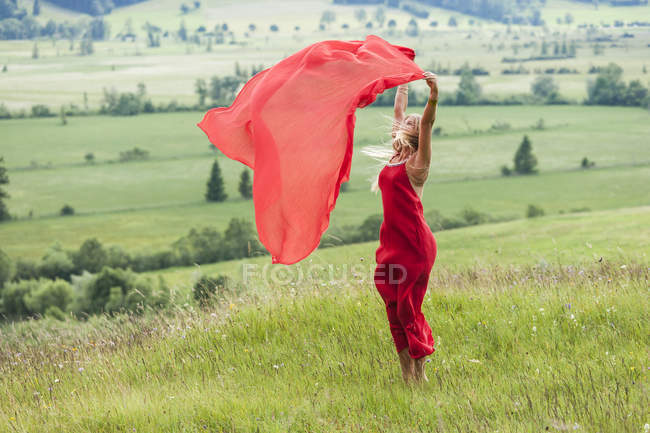 Femme en robe rouge debout sur une prairie tenant un tissu rouge — Photo de stock