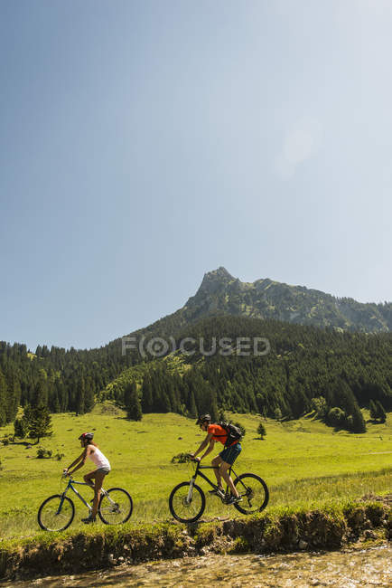 Áustria, Tirol, Vale de Tannheim, jovem casal andando de bicicleta de montanha à beira-rio — Fotografia de Stock