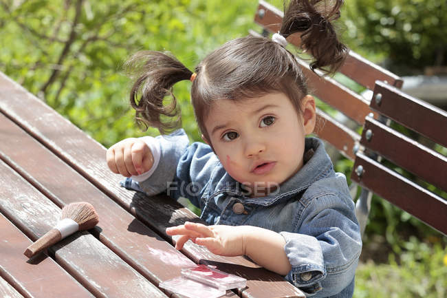Portrait of little girl sitting at garden table with make-up brush and rouge — Stock Photo