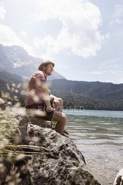 Alemania, Baviera, Eibsee, hombre sonriente en Lederhosen sentado a orillas del lago - foto de stock