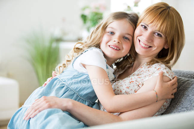 Madre e hija sentadas en el sofá, abrazadas - foto de stock