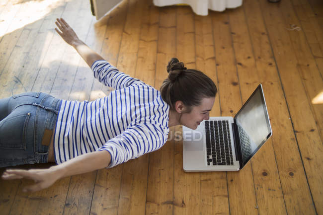 Jeune femme à la maison couchée sur le sol en bois avec ordinateur portable, faisant semblant de voler — Photo de stock