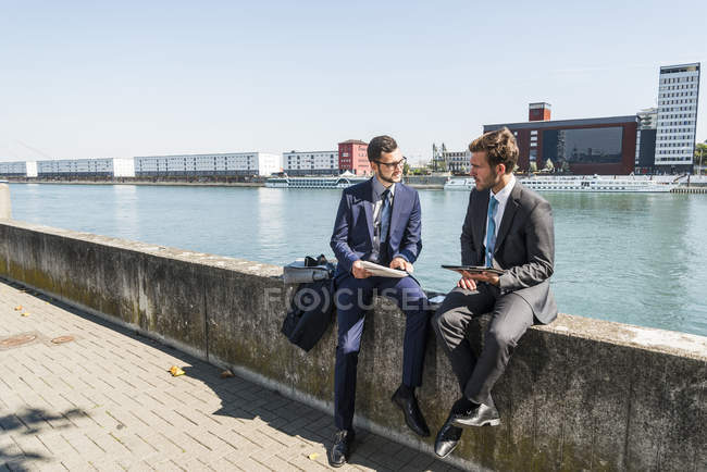 Dos jóvenes empresarios sentados en la pared junto al río, trabajando - foto de stock