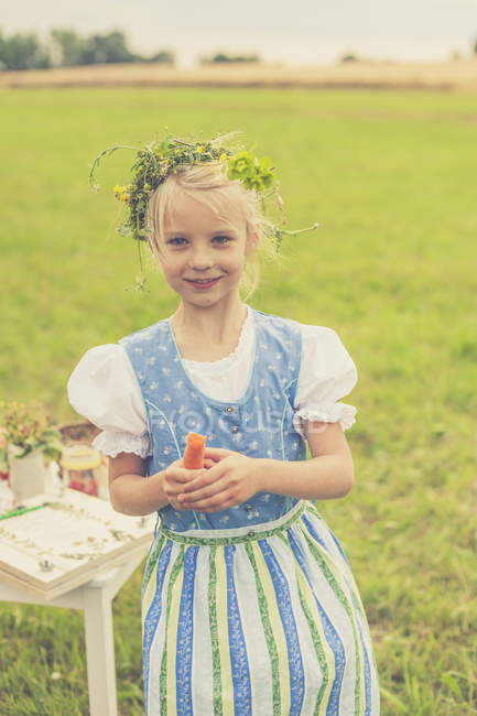 Germany, Saxony, portrait of smiling girl with floral wreath and dirndl holding carrot in her hands — Stock Photo