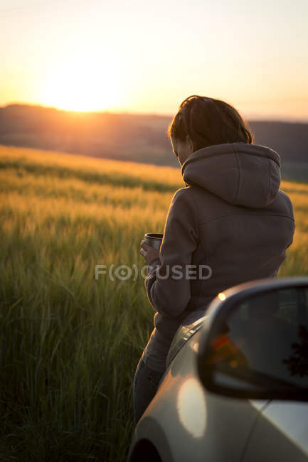 Mulher inclinada no carro na frente de um campo ao nascer do sol — Fotografia de Stock