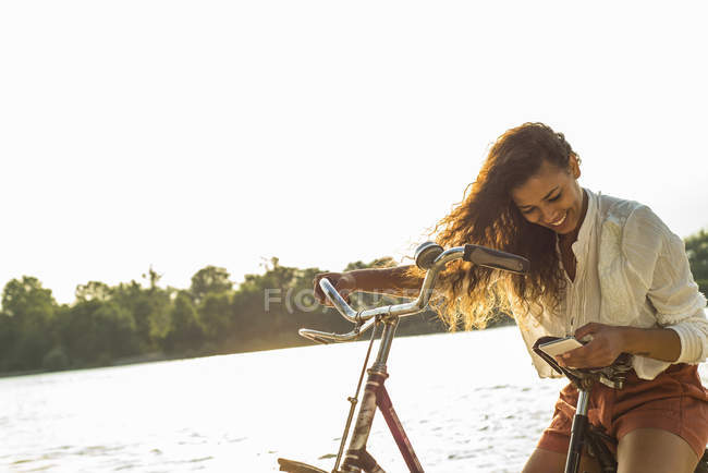 Mujer joven sonriente con bicicleta y teléfono celular a la orilla del río - foto de stock