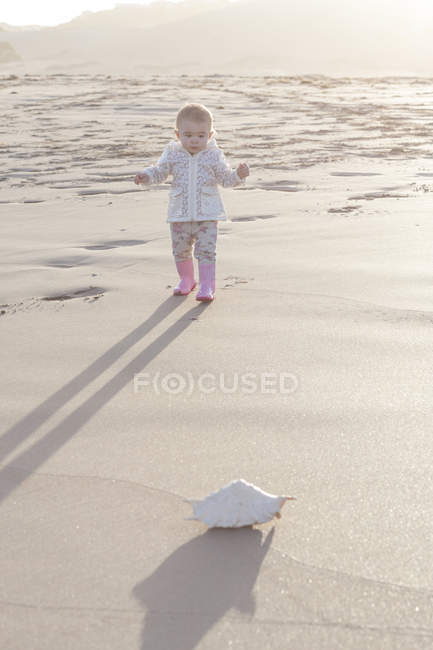 Babymädchen steht am Strand und blickt auf eine Muschel — Stockfoto