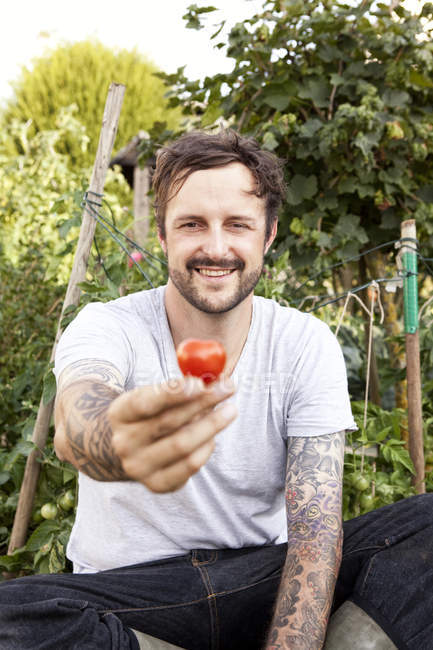 Ritratto di uomo sorridente con tatuaggi sulle braccia seduto nel giardino che tiene il pomodoro — Foto stock