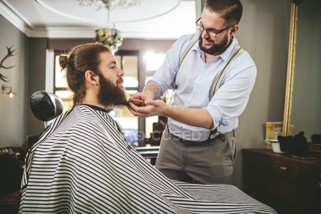 Barber brushing beard of a customer in barbershop — cape, smiling ...
