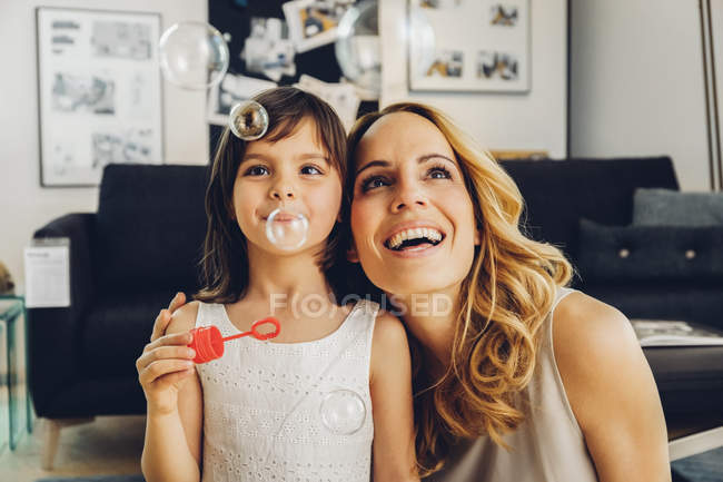 Mère heureuse avec sa fille à la maison soufflant des bulles de savon — Photo de stock