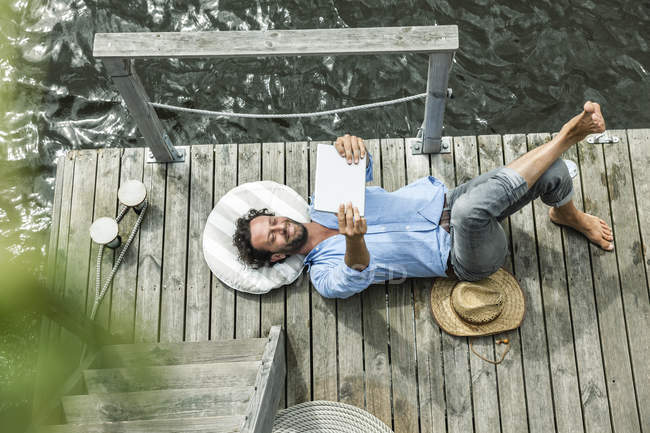 Mann liegt mit digitalem Tablet auf Bahnsteig am Wasser — Stockfoto