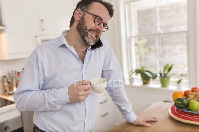 Uomo in piedi in cucina con coppa di telefonare con smartphone — Foto stock